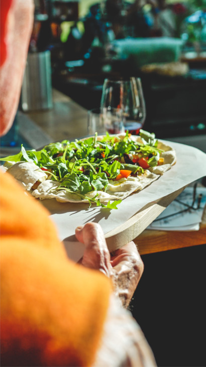 Vegan tarte flamb&eacute;e with tomato sauce, bell pepper, olives, and arugula, drizzled with olive oil, served on a wooden board.