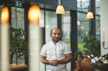 John Kasraei, tenant and chef, standing in the winter garden of the Element restaurant, wearing a chef&rsquo;s jacket with hands clasped in front.