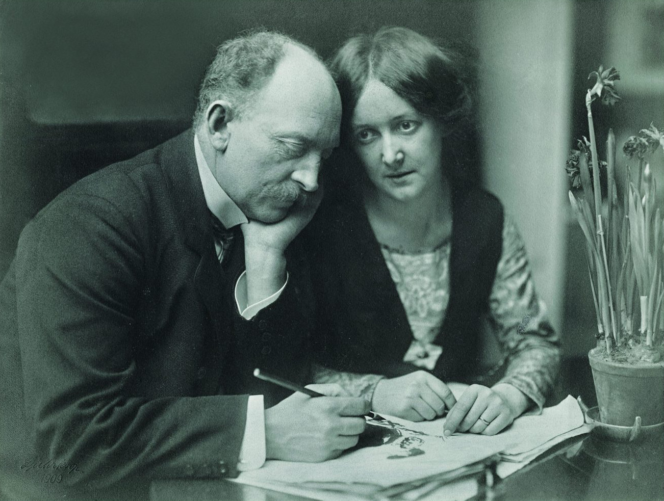 Historical photo of Ada and Emil Nolde, sitting side by side at a table; Emil Nolde with a fountain pen and papers in front of him.