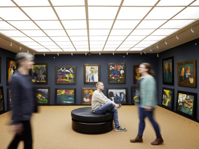 Impression aus dem Bildersaal des Nolde-Hauses mit dunkelblauen W&auml;nden, dichter H&auml;ngung von Werken Emil Noldes und Besuchern in Bewegung.